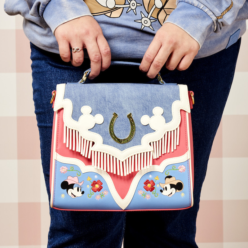 Close up shot of person holding the Loungefly Western Mickey and Minnie Fringe Crossbody Bag in front of the camera against a soft pink checkered background